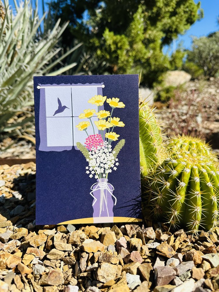 Art print by MidSun Designs of a vase with a wildflower bouquet in front of a window with a hummingbird. Art print placed outside by cacti.